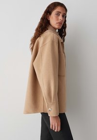Veste en mélange de laine beige avec une coupe décontractée, un poignet boutonné et une grande poche poitrine. Texture lisse et détails de large col.