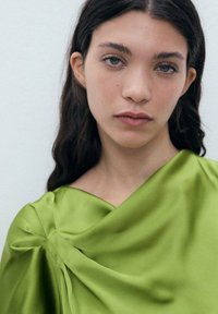 Blouse en satin vert avec un design drapé et un détail en nœud à l'épaule. Sa texture lisse et son éclat doux rehaussent son apparence.
