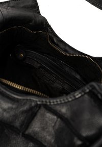 Close-up van een open zwart leren tas met zichtbare ritszak en een reliëflabel met de tekst "RE:DESIGNED EST. 2005" aan de binnenkant.