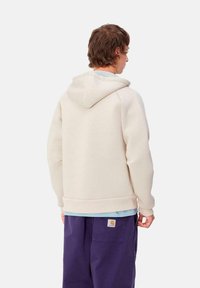 Sudadera de color crema con capucha ajustable, puños y dobladillo acanalados. Tela suave y texturizada, llevada sobre una camisa de color azul claro; combinada con pantalones morados.