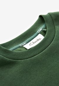Felpa verde scuro con scollo rotondo. Presenta una consistenza del tessuto liscia e un'etichetta del marchio visibile con la scritta "Clarks" in bianco.