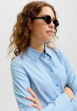 Woman with curly brown hair wearing round black sunglasses and a blue striped button-up shirt, arms crossed, facing right.