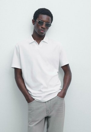 Giovane uomo con occhiali da sole scuri, polo bianca a maniche corte e pantaloni grigio chiaro, in piedi contro un muro bianco con le mani in tasca.