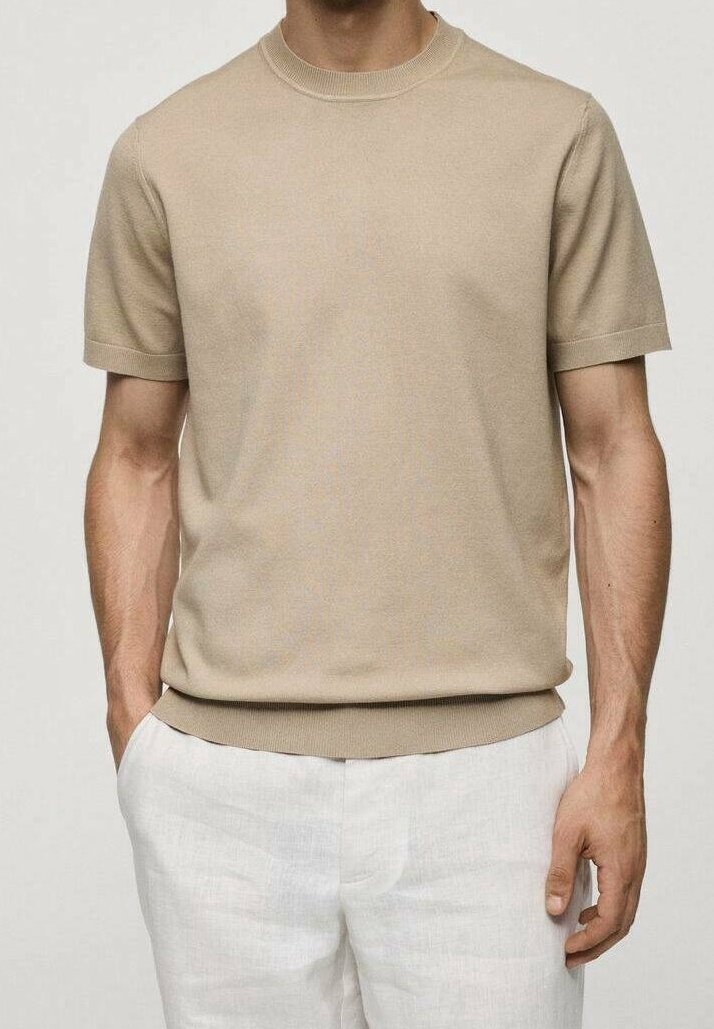 Homme portant une chemise en maille beige à manches courtes et un pantalon en lin blanc, debout avec une main dans la poche devant un fond uni.
