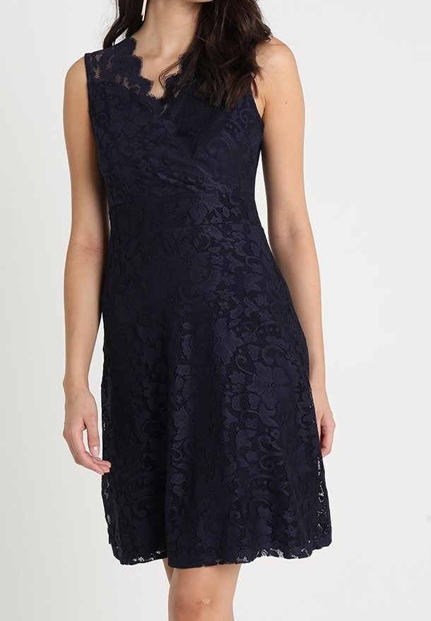 Femme portant une robe en dentelle bleu marine sans manches, longueur genou, avec un décolleté en V festonné et un motif floral.