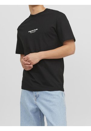 Personne portant un t-shirt noir avec un texte blanc "Originals Studio" et un jean bleu clair, debout devant un fond gris clair uni.