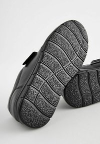 Chaussures noires avec une semelle en caoutchouc texturé présentant un motif de bande de roulement. Le matériau supérieur est lisse, avec une sangle velcro d'un côté.