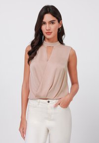 Top sin mangas color beige con cuello alto simulado, con un corte en forma de llave en el centro y un diseño drapeado, combinado con jeans blancos de cintura alta.