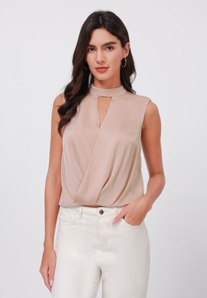 Beige mouwloos top met een opstaande kraag, voorzien van een centrale sleutelgatuitsparing en een gedrapeerd ontwerp, gecombineerd met witte high-waisted jeans.