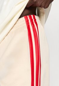 Beige Sporthose mit roten und weißen vertikalen Streifen an den Seiten, aus weichem Material und mit elastischem Bund.