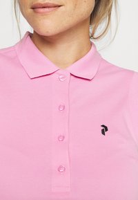 Różowa koszulka polo z kołnierzykiem i czarnym logo. Posiada sześć guzików z przodu i fakturowaną tkaninę.