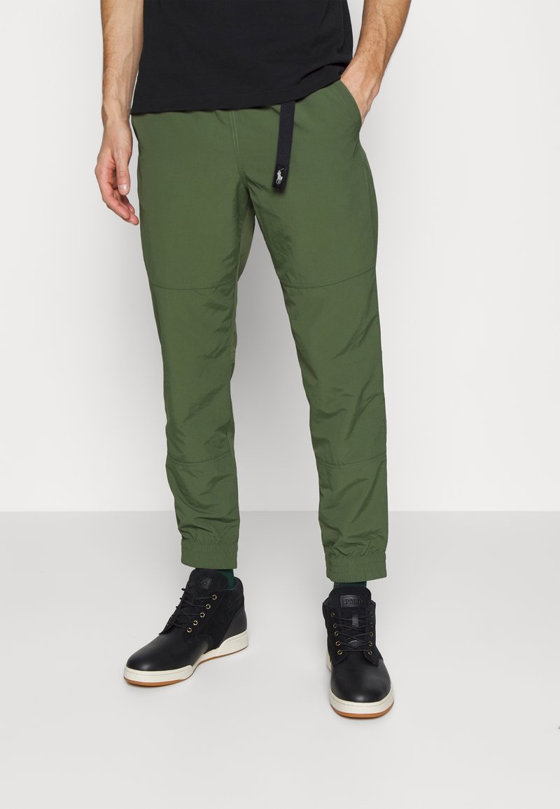 Homme portant un pantalon de jogging vert fuselé avec des poignets élastiques et des baskets montantes noires, debout devant un fond clair.