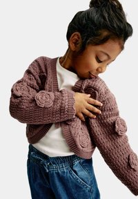 Stickad cardigan i mauve med texturerat mönster och blomsterdetaljer på ärmarna. Bärs över en vit topp, tillsammans med jeansbyxor.