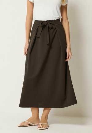 Kvinde iført en mørkebrun midi-nederdel med højtaljet, med bindebånd, hvid top med korte ærmer og beige flade sandaler.