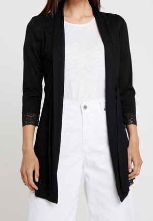 Vrouw draagt een zwart vest met open voorkant en kanten geruite mouwen over een wit T-shirt en witte broek met hoge taille.