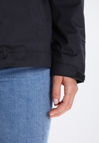Main détendue à côté d'un jean denim bleu, portant une veste noire avec un poignet Velcro sur fond clair.