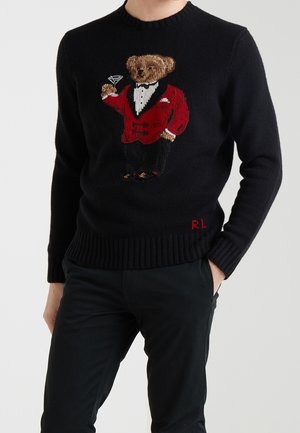 Mies pukeutuneena mustaan villapaitaan, jossa on punapukuinen nallekarhu ja martini-lasi kädessä, yhdistettynä mustiin housuihin.