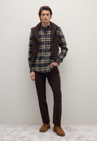 Gilet trapuntato marrone sopra una camicia a quadri blu e verde con bottoni, pantaloni di velluto a coste marrone scuro e scarpe in camoscio marrone. Design semplice e strutturato.