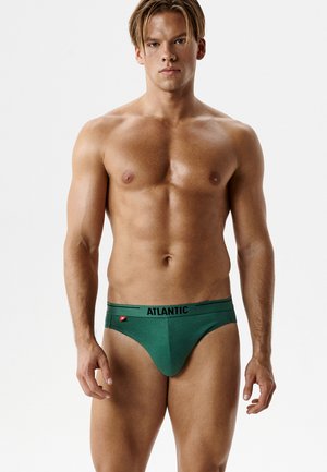 Młody muskularny mężczyzna stojący, ubrany w zielone slipy z paskiem oznaczonym „ATLANTIC” na prostym białym tle.