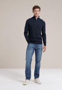 Marinefarbener Zip-Kragen-Pullover aus glattem Strickstoff, kombiniert mit hellblauen Jeans und weißen Sneakers. Minimalistisches Design und lässige Passform.