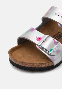 Sandal s dva remena sa srebrnim sintetičkim gornjim dijelom koji sadrži šarene točkice. Korkana unutrašnjost, crna gumena potplat i podesiva metalna kopča.