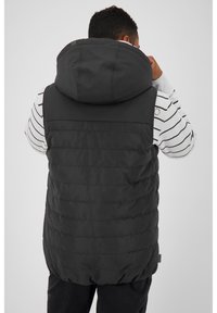 Zwarte gewatteerde vest met een grote capuchon, met een geborduurde textuur en een relaxte fit. Draag het over een wit met zwarte gestreepte lange mouw top.