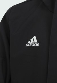 Veste adidas noire avec un logo blanc sur la poitrine. Le matériau semble lisse et synthétique avec un design moderne et ajusté.