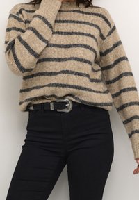 Beżowy sweter z dzianiny z czarnymi poziomymi paskami, okrągłym dekoltem i długimi rękawami. Noszony z czarnymi dżinsami i ozdobnym paskiem.