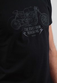 Črna majica s potiskom motocikla z zapletenimi črtami. Besedilo pravi "STROJ SLOBODE" z bliski.