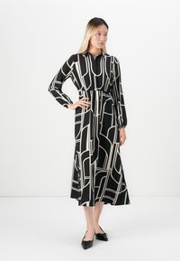 Femme se tenant debout avec une main sur la hanche, portant une longue robe noire avec des lignes géométriques blanches et des ballerines noires à bout pointu.
