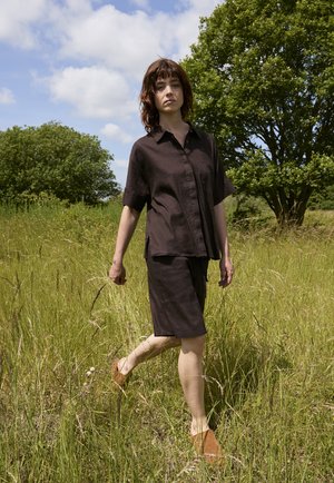 Frau, die durch hohes Gras geht, trägt ein schwarzes kurzärmeliges Shirt und Shorts, mit braunen Slippern, unter teilweise bewölktem Himmel.