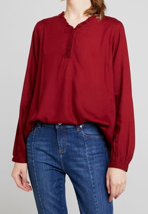 Femme portant un chemisier rouge à manches longues avec un col en V à volants et un jean bleu, debout devant un fond gris clair.