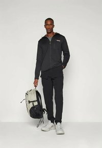 Black zippered hooded jacket over a gray shirt, paired with black pants. The model holds a gray and black backpack. Footwear is light gray.