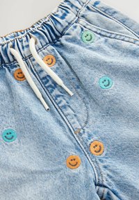 Gaiši zilas džinsu šorti ar elastīgu jostu. Tajos ir daudzkrāsains smaidošs sejiņas izšuvums un baltas aukliņas.