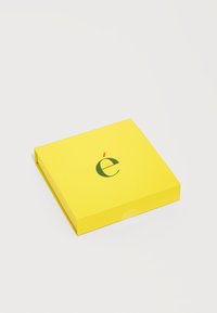 Boîte carrée jaune vif avec une lettre "é" accentuée en vert et rouge, centrée en haut, sur un fond blanc uni.