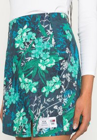 Närbild av en blommönstrad kjol med gröna och vita blommor på mörkblått tyg, med en slits framtill och Tommy Jeans -etikett.