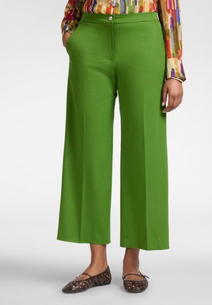 Person trägt hoch taillierte, weit geschnittene grüne Hosen kombiniert mit braunen geflochtenen Ballerinas und einem mehrfarbigen Langarmhemd.