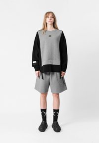 Sudadera gris con mangas negras, con un logo. Combinada con pantalones cortos grises, detalle de cadena, y calcetines negros con un patrón blanco. Calzado negro.