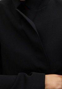Chaqueta negra con un cuello alto, con una solapa asimétrica doblada, textura suave y mangas largas. La tela parece ligera.