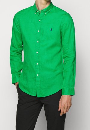 Mann trägt ein leuchtend grünes, langärmeliges Hemd mit Knöpfen und einem kleinen dunklen Logo auf der Brust, kombiniert mit einer schwarzen Hose.