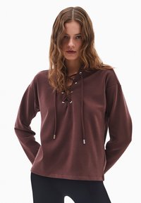 Brązowy sweter z sznurkiem z przodu i srebrnym wykończeniem. Posiada długie rękawy i luźny krój, wykonany z miękkiego, fakturowanego materiału.