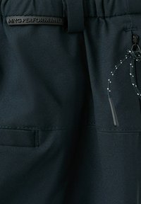 Gros plan sur un pantalon de sport noir montrant une taille élastique, un cordon de serrage avec des points réfléchissants, une poche zippée et une étiquette « MNG PERFORMANCE ».