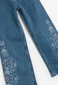 Vaqueros de mezclilla en azul oscuro con bordados florales en blanco y rojo a lo largo de los extremos exteriores. Diseño de pierna recta con una altura media.