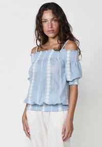 Blusa azzurro chiaro a spalle scoperte realizzata in tessuto testurizzato, con motivi ricamati bianchi e maniche a sbuffo, e orlo arricciato.