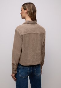 Femme de dos portant une chemise en velours côtelé beige à boutons et un jean en denim bleu, sur un fond clair uni.