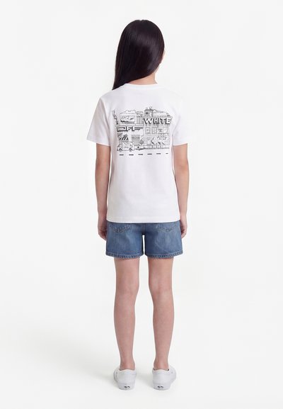 Παιδί που στέκεται πλάτη, φορώντας λευκό μπλουζάκι με μαύρο αστικό γκράφιτι και μπλε τζιν σορτς με λευκά αθλητικά παπούτσια.