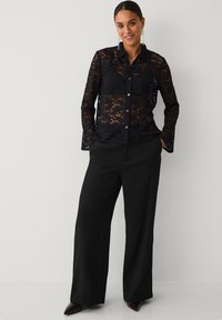 Schwarzes Spitzenhemd mit Knopfleiste und figurbetontem Schnitt. Lange Ärmel aus Spitze und ein Kragen. Kombiniert mit weiten schwarzen Hosen und spitzen Schuhen.