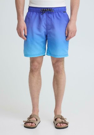 Mand iført blå gradient badeshorts med sort og hvid snor, stående barfodet i beige sandaler mod en ensfarvet baggrund.