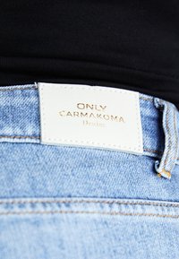 Jeans de denim azul claro com uma etiqueta de couro branco com letras douradas, mostrando o nome da marca "ONLY CARMAKOMA" e "Denim".