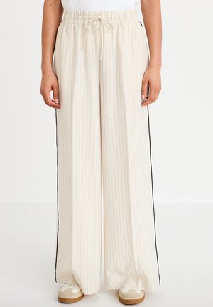 Kremfarvede bukser med vide ben med tynde lodrette striber og sorte side-striber, elastisk talje med snøre, båret med beige sneakers.